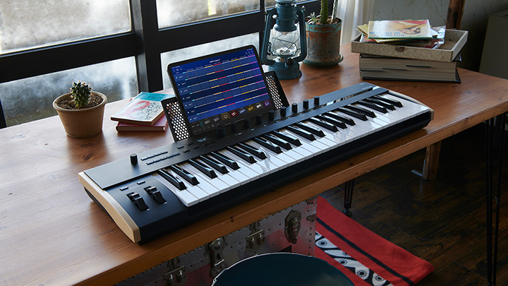 A digital keyboard synthesizer sits on a wooden desk with a tablet displaying music software on its stand. A small potted cactus, lantern, books, and other items decorate the workspace near a window.