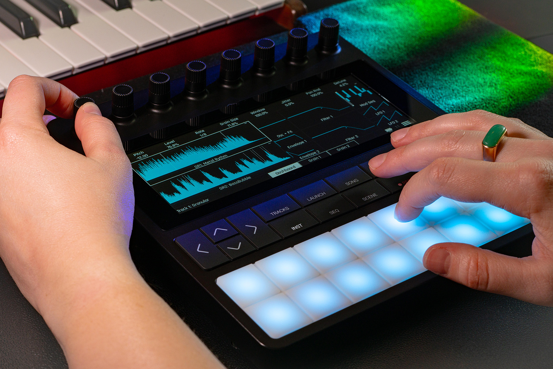 A person uses a music production controller with a touchscreen, knobs, and glowing blue pads, next to a keyboard. A green ring is visible on one finger, and colorful lights shine on the surface.