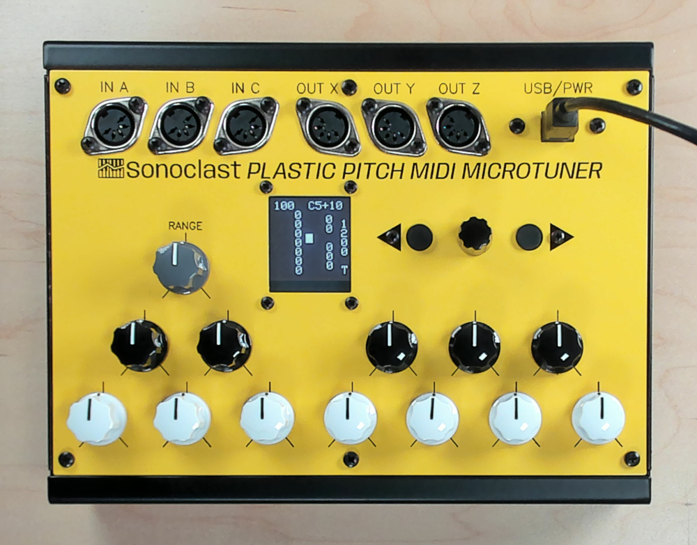 A yellow Sonoclast Plastic Pitch MIDI Microtuner with multiple knobs, switches, a digital display, connection ports labeled IN/OUT, and a USB/PWR port on a light wood surface.