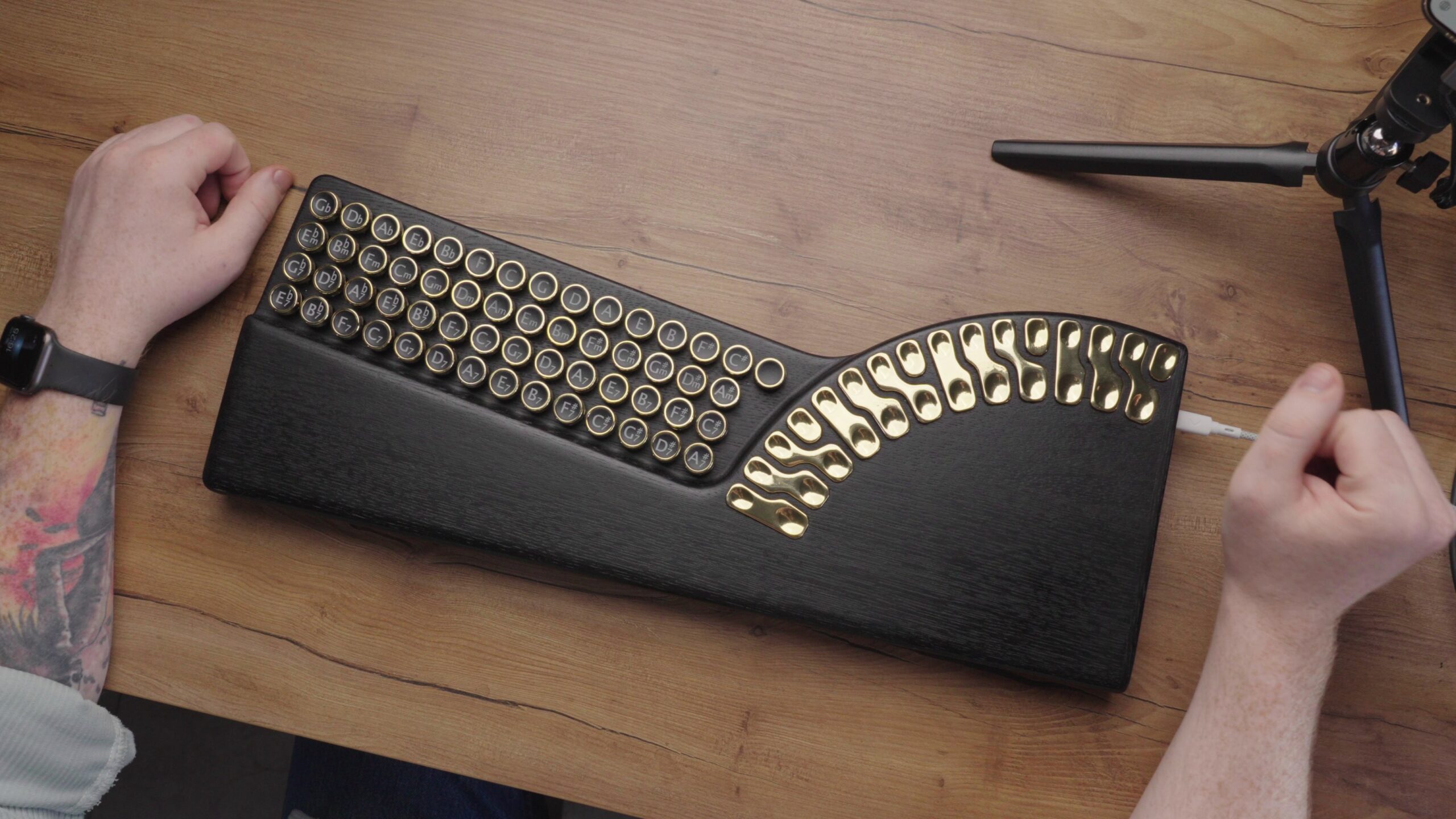 A person’s hands rest near a unique black and gold keyboard with round keys and a curved patterned metal section, placed on a wooden desk beside a tripod.