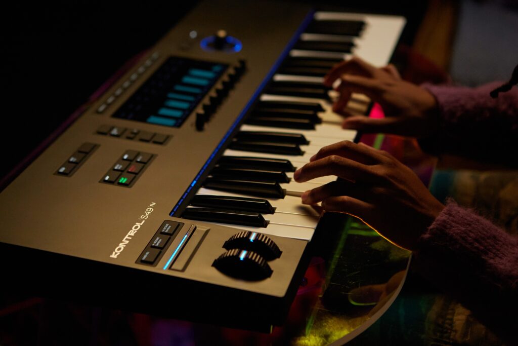 A person is playing a black electronic keyboard with illuminated buttons and displays in a dimly lit room. Their hands are on the keys, and the instrument is labeled KONTROL S49 MK2.