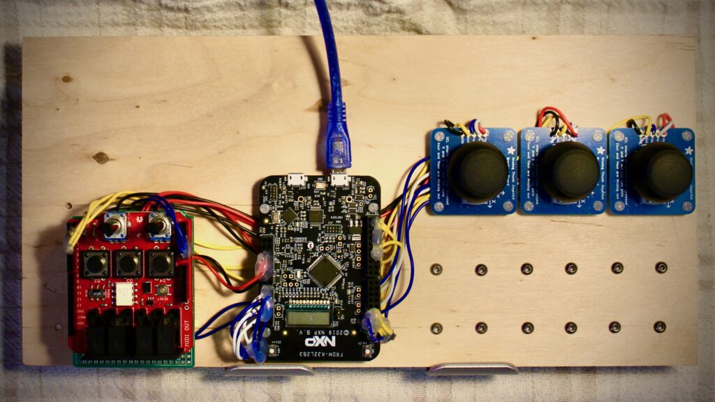 A wooden board with a circuit setup: a red module with buttons on the left, a central black microcontroller connected by USB, and three blue joystick modules on the right, all wired together.