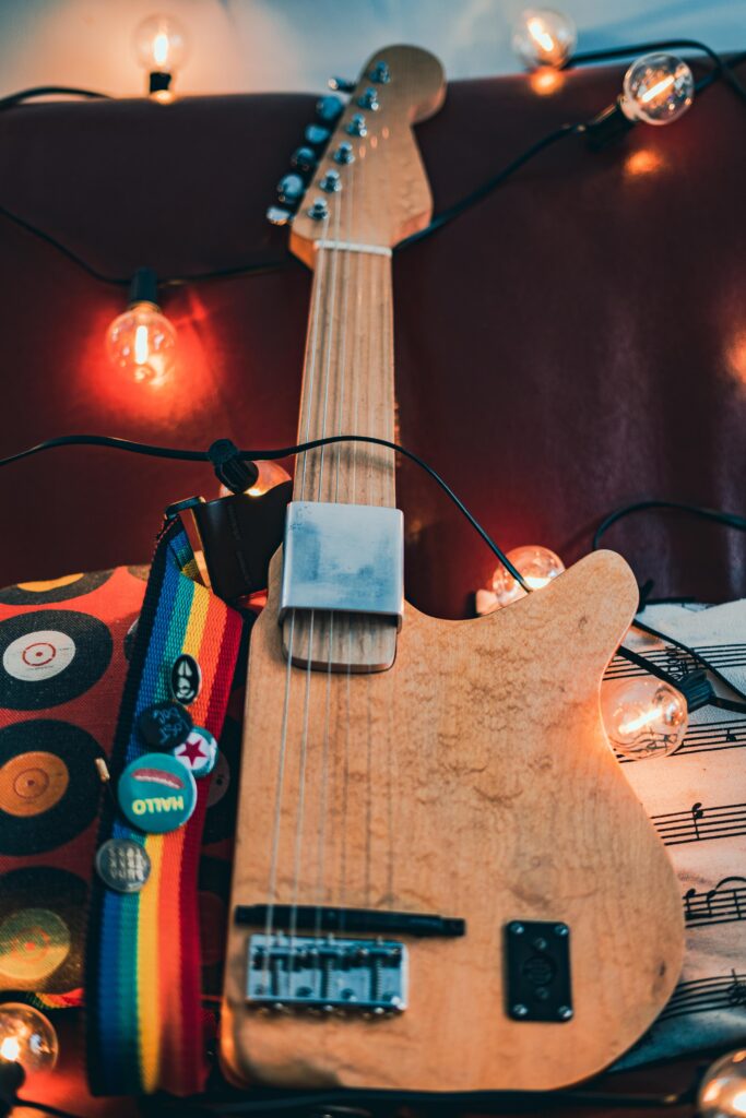 A wooden electric guitar lies on a red surface, surrounded by glowing string lights, a colorful rainbow strap with pins, and a pillow featuring musical notes.