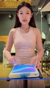 A woman in a light pink sleeveless dress stands indoors, playing an electronic musical instrument with a glowing, multicolored touchpad. Studio lighting and recording equipment are visible in the background.