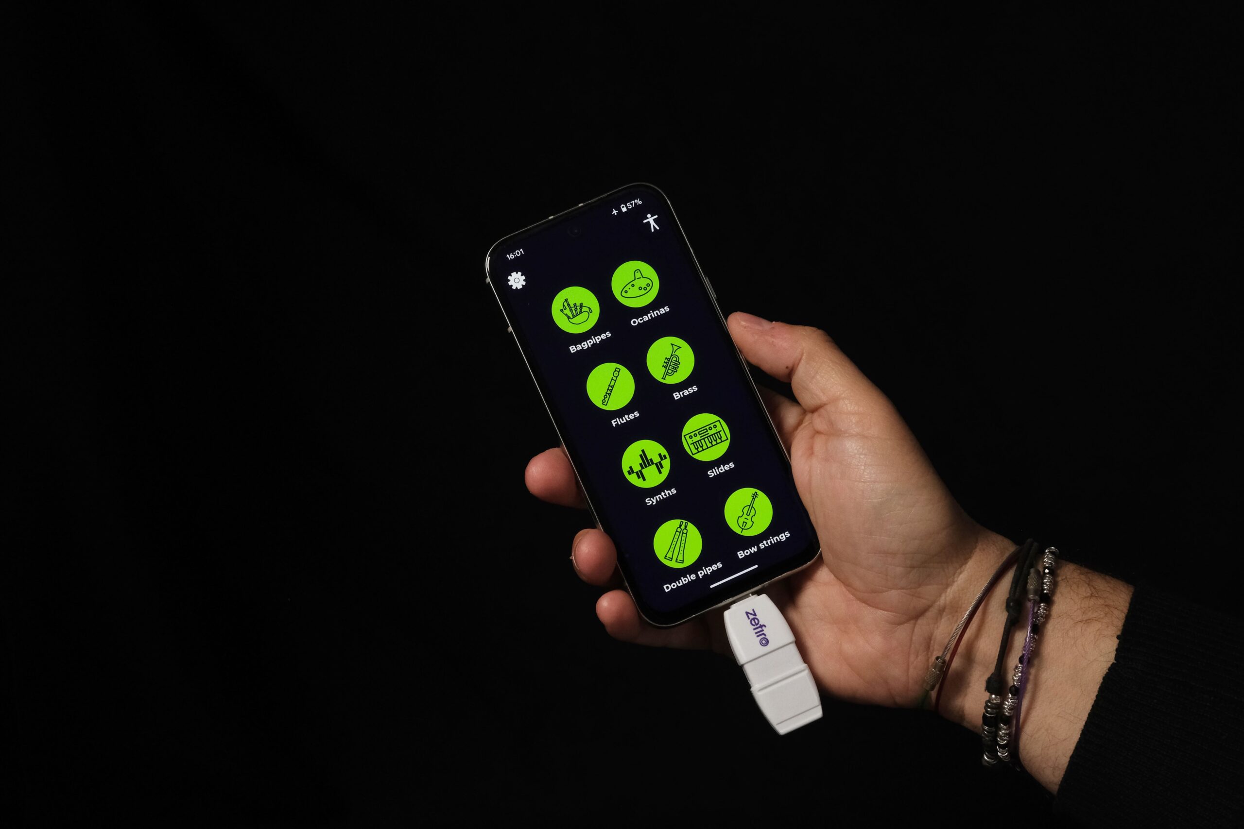 A hand holds a smartphone displaying a screen with green circular app icons and Russian text. A white device is plugged into the phone’s charging port. The background is black.