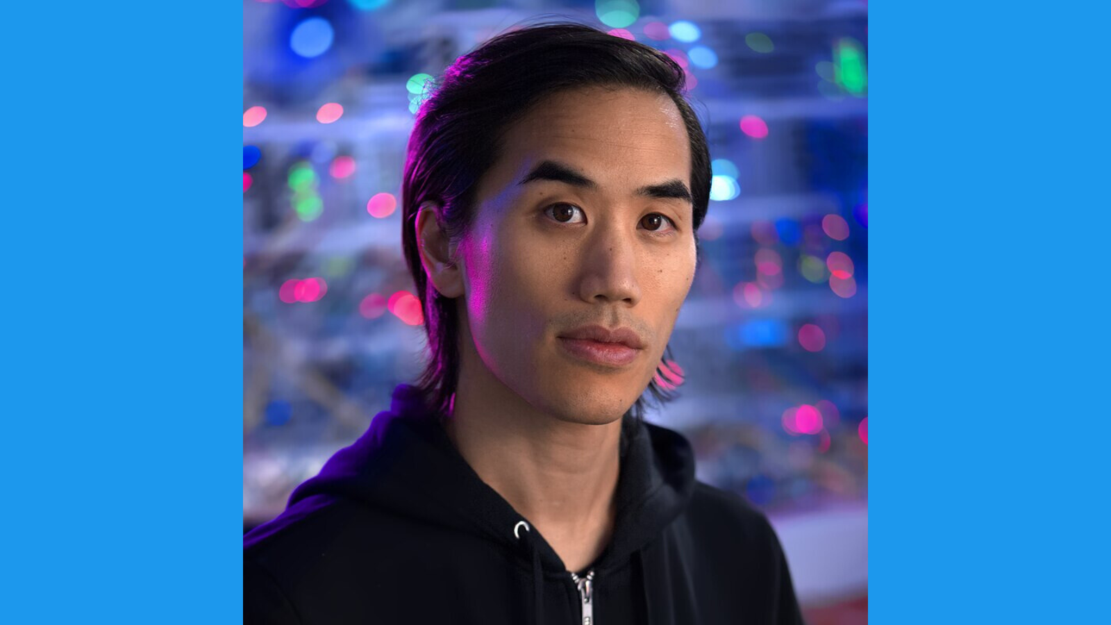 Andrew Huang with long dark hair, wearing a black hoodie, looks at the camera with a neutral expression. Colorful, out-of-focus lights create a vibrant background. The image is framed by blue bars on each side.