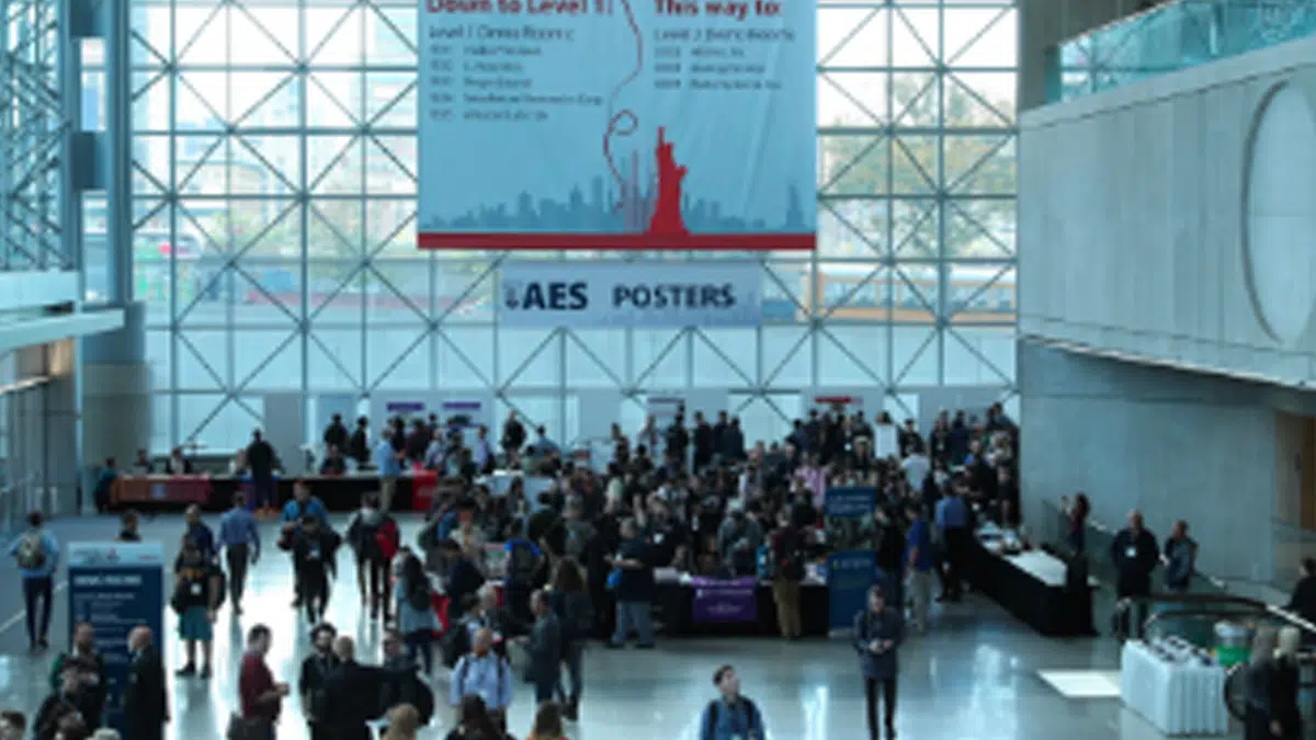 A large crowd gathers in a spacious convention center lobby with tall windows. A banner with a red Statue of Liberty graphic hangs above booths and people. Sunlight streams inside, creating a bright atmosphere.