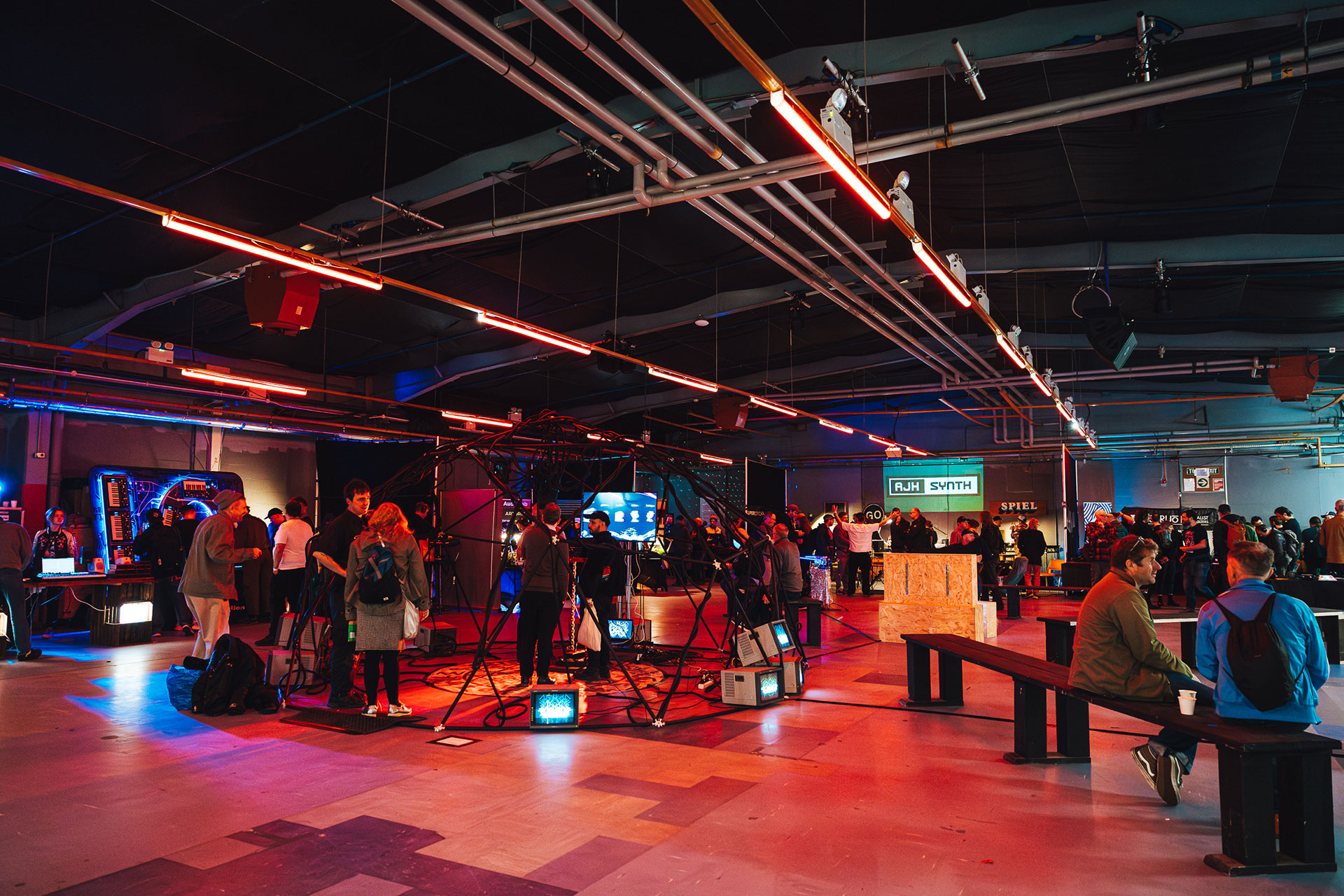 A large, colorful indoor event space filled with people interacting at various tech booths and displays, under red and blue lighting, with benches and electronic devices scattered throughout.