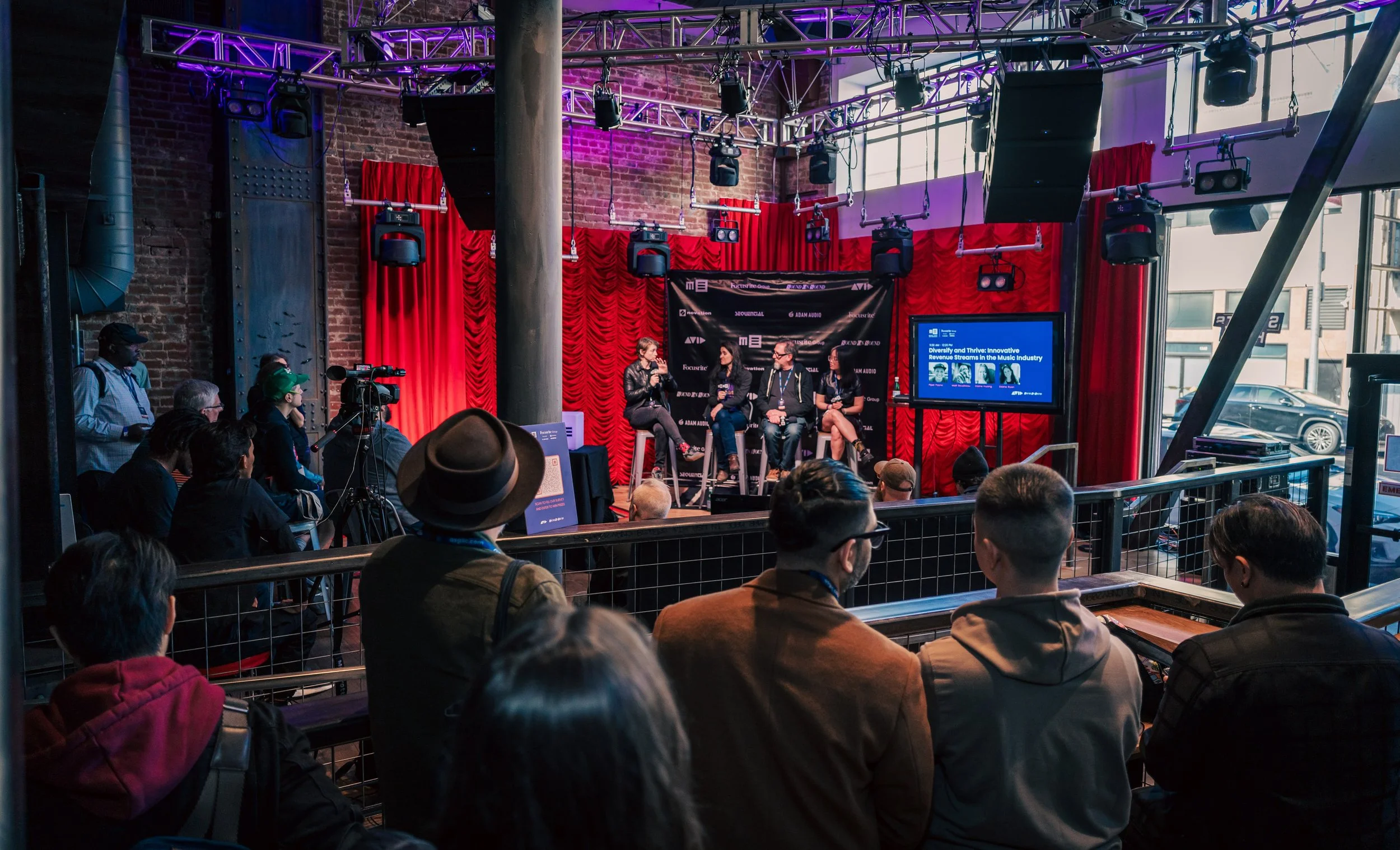 A panel discussion is taking place onstage in front of an audience in an indoor venue with red curtains, stage lighting, and a screen displaying information. People are seated and standing, watching the speakers.