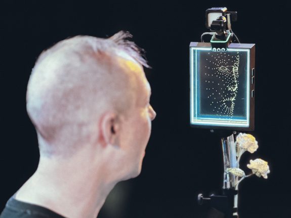 A person looks at a digital screen displaying a dotted, abstract facial outline. The screen is mounted on a stand with a camera above it, and two white flowers are attached to the stand below the screen.