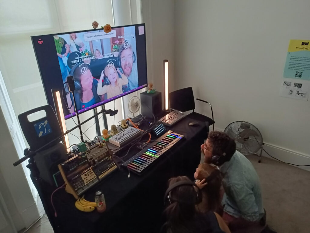 A man and two children wearing headphones sit in front of an electronic music workstation with colorful keys, audio equipment, and a large screen showing a group selfie with augmented reality filters.