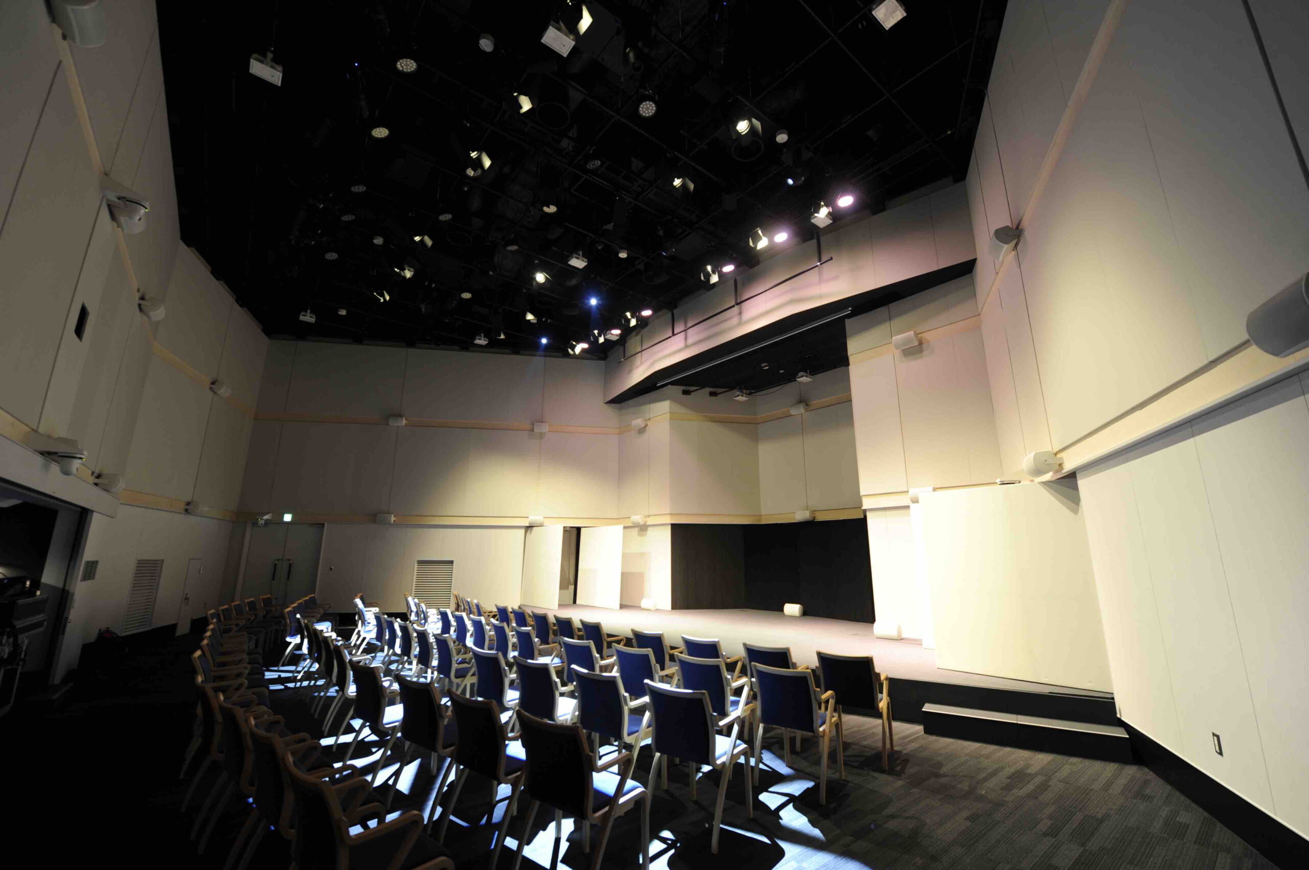 A modern, empty auditorium with rows of chairs facing a well-lit stage. The walls are white and the ceiling is equipped with stage lighting. No people are present, and the space appears ready for an event or performance.
