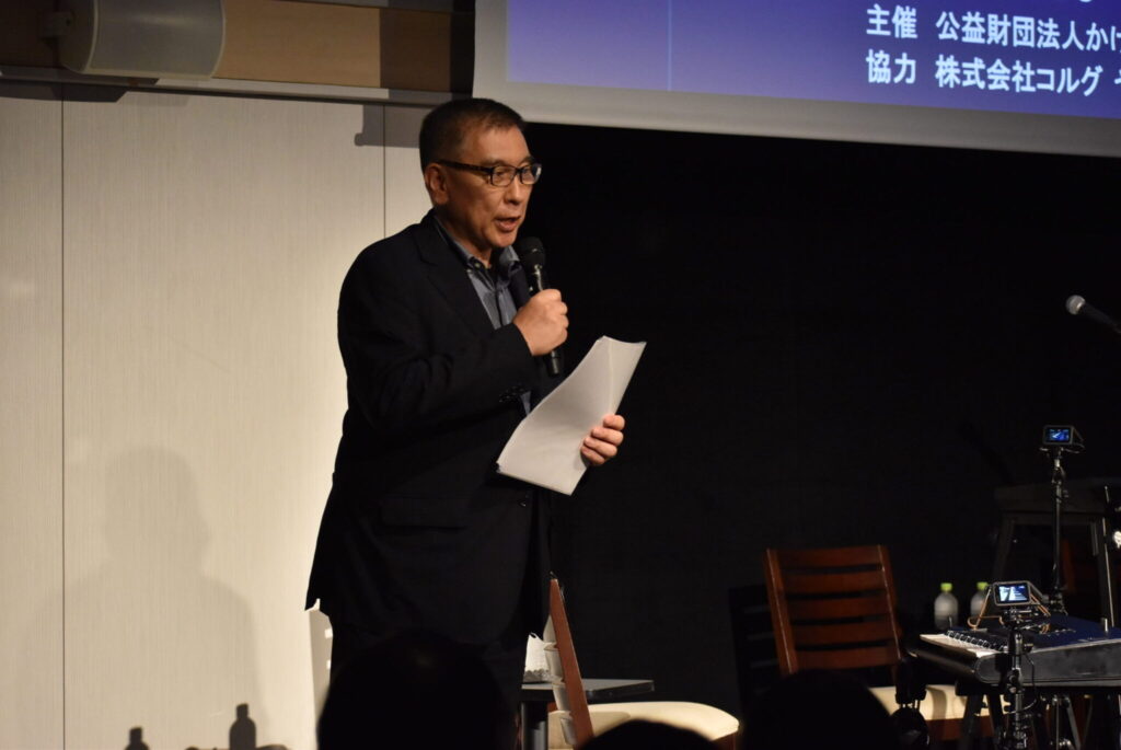 A man in a dark suit and glasses speaks into a microphone while holding papers on a dimly lit stage, with a projection screen and chairs in the background. Text in Japanese is partially visible on the screen above him.