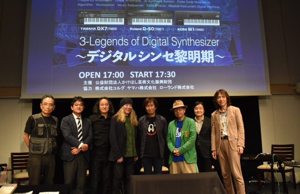 Eight people stand on stage in front of a large screen displaying images of synthesizers and text about 3-Legends of Digital Synthesizer. The group is posing for a photo at a music-related event.