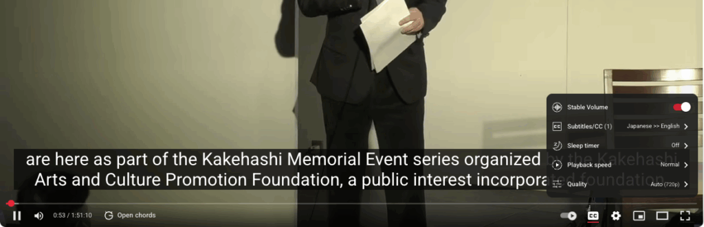 A person in a suit stands on stage holding papers, partially visible, with YouTube video controls and English subtitles displayed at the bottom of the screen. A pop-up menu shows playback and subtitle options on the right.