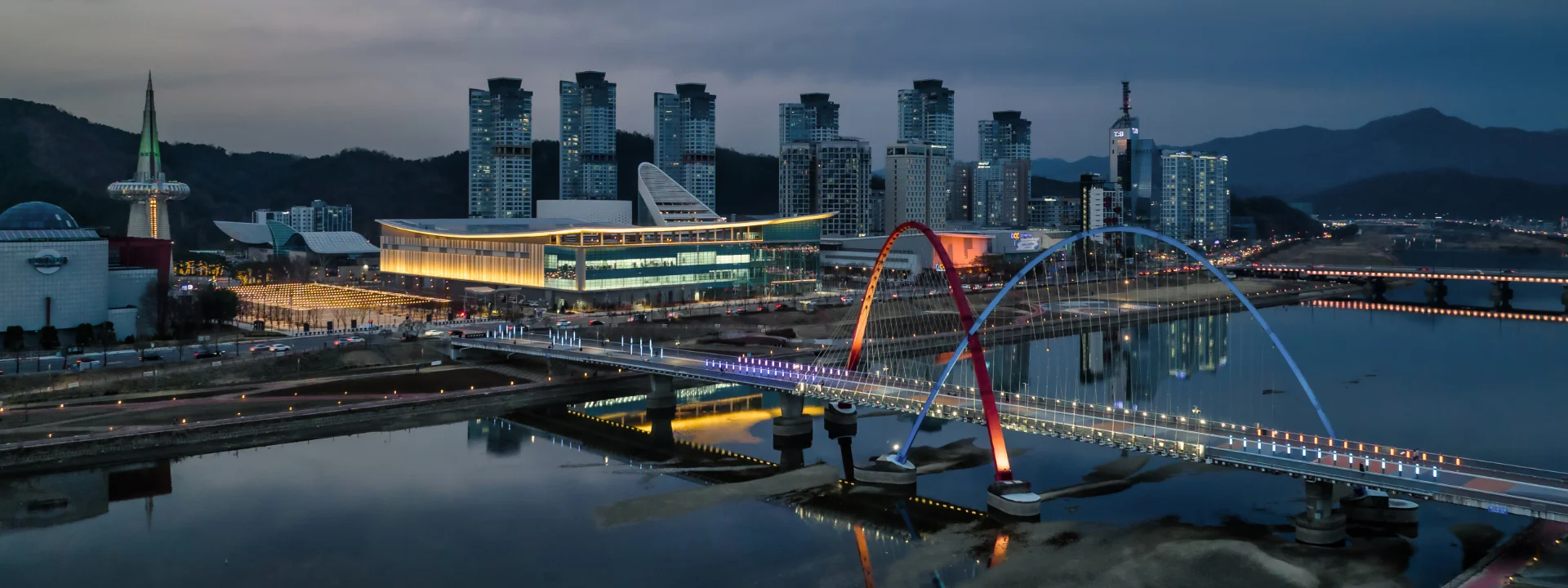 A cityscape at dusk shows modern high-rise buildings, a brightly lit convention center, and a colorful arched bridge over a calm river, with reflections of the lights on the water. Hills and a tower are visible in the background.