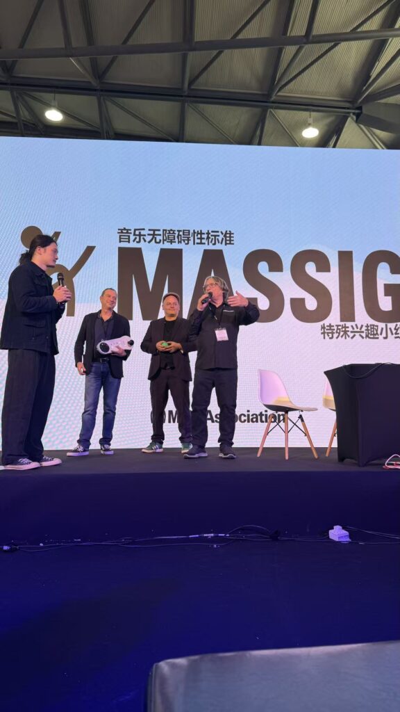 Four people stand on a stage speaking into microphones in front of a large screen displaying the word MASSIG and text in Chinese characters. One person gestures with their hand while another holds a small object.