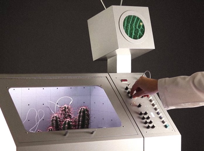 A person in a lab coat adjusts knobs on a futuristic control panel with a cactus inside a glass compartment and a monitor above displaying a green pattern.