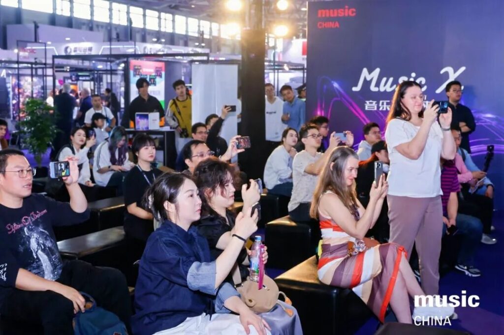 A diverse audience sits and stands at an indoor event, many holding up phones to take photos. A large music China sign is visible in the background. The atmosphere is lively and engaged.