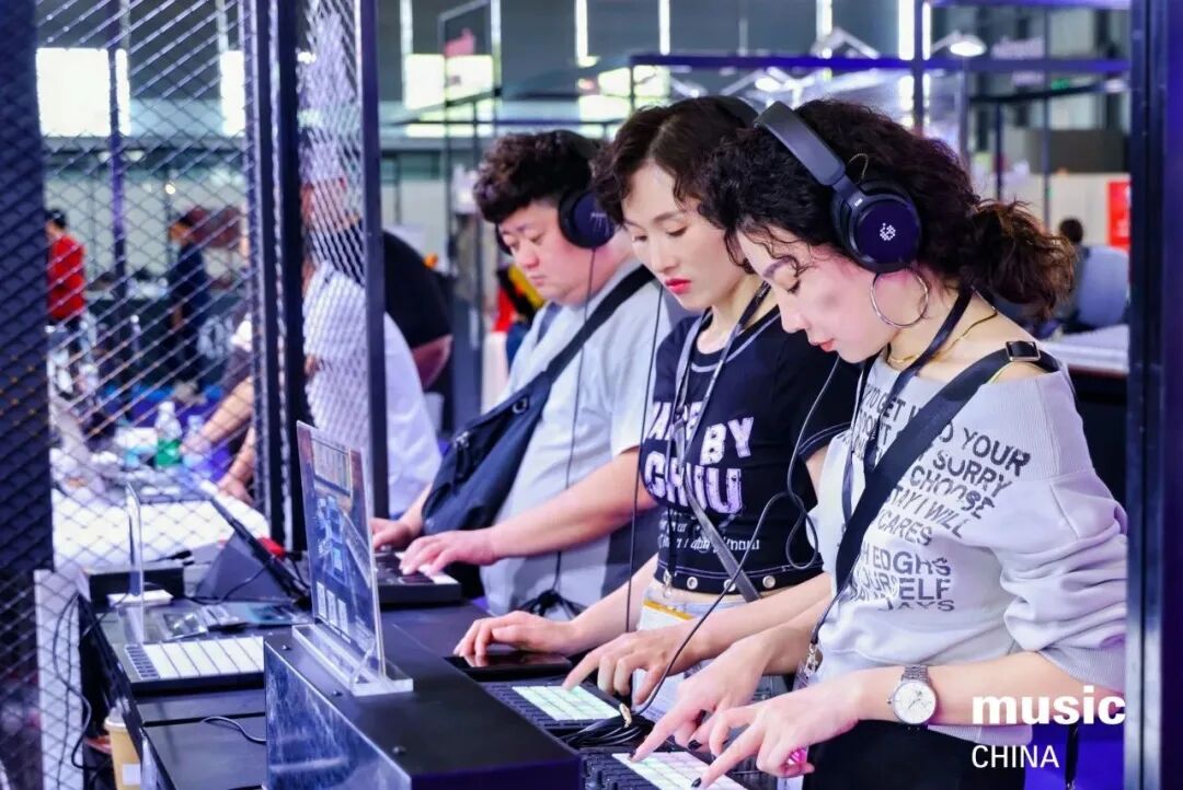Three young people wearing headphones use electronic music equipment at a booth. They are focused on creating music, with laptops and controllers in front of them. The scene appears to be at a music expo or event.
