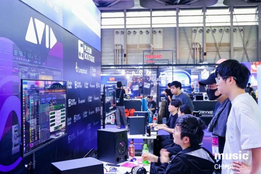 A group of people gathers around a large screen displaying audio software at a technology or music expo, with various booths and electronic equipment visible in the background.