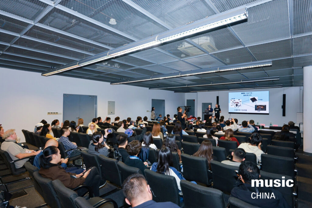 A group of people sit in rows of seats, attentively watching a presentation in a modern conference room. A speaker stands at the front near a screen displaying slides. The words music CHINA are visible in the lower right corner.