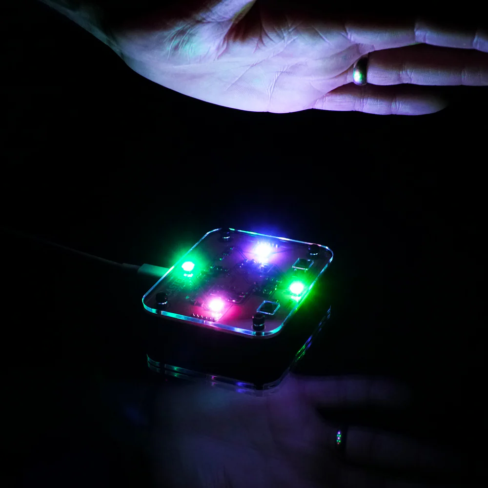 A small electronic device with multicolored LED lights glows in the dark, while two hands hover above and below it, partially illuminated by the lights.