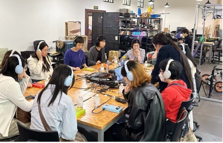 A group of people wearing headphones sit around a large table covered with audio equipment and laptops, engaged in discussion in a modern, well-lit workspace.