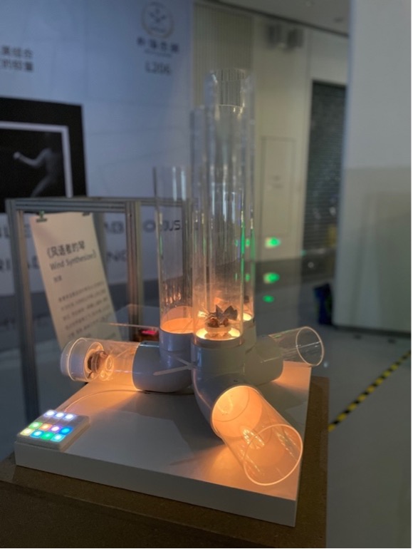 A modern wind synthesizer machine on display, with illuminated cylinders, transparent tubes, and a digital control panel, set in a well-lit indoor exhibition space.