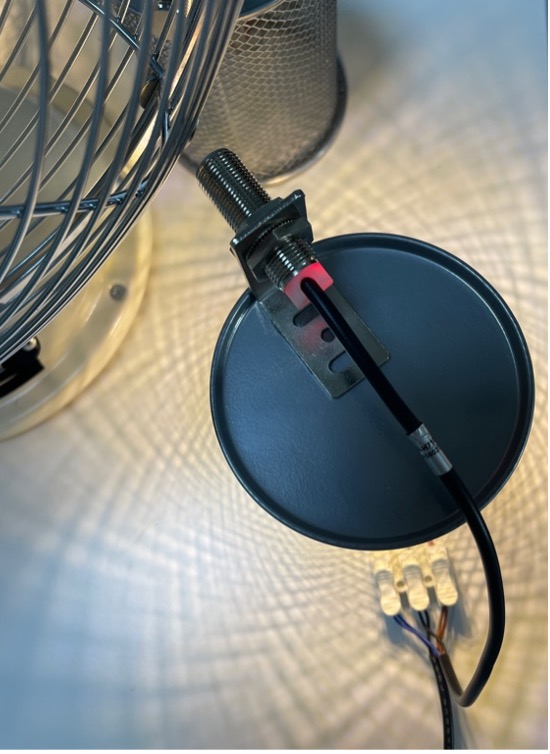 Close-up of a metallic sensor mounted on a round, dark surface with a black cable attached. A wire mesh and diffused lighting create a patterned background.