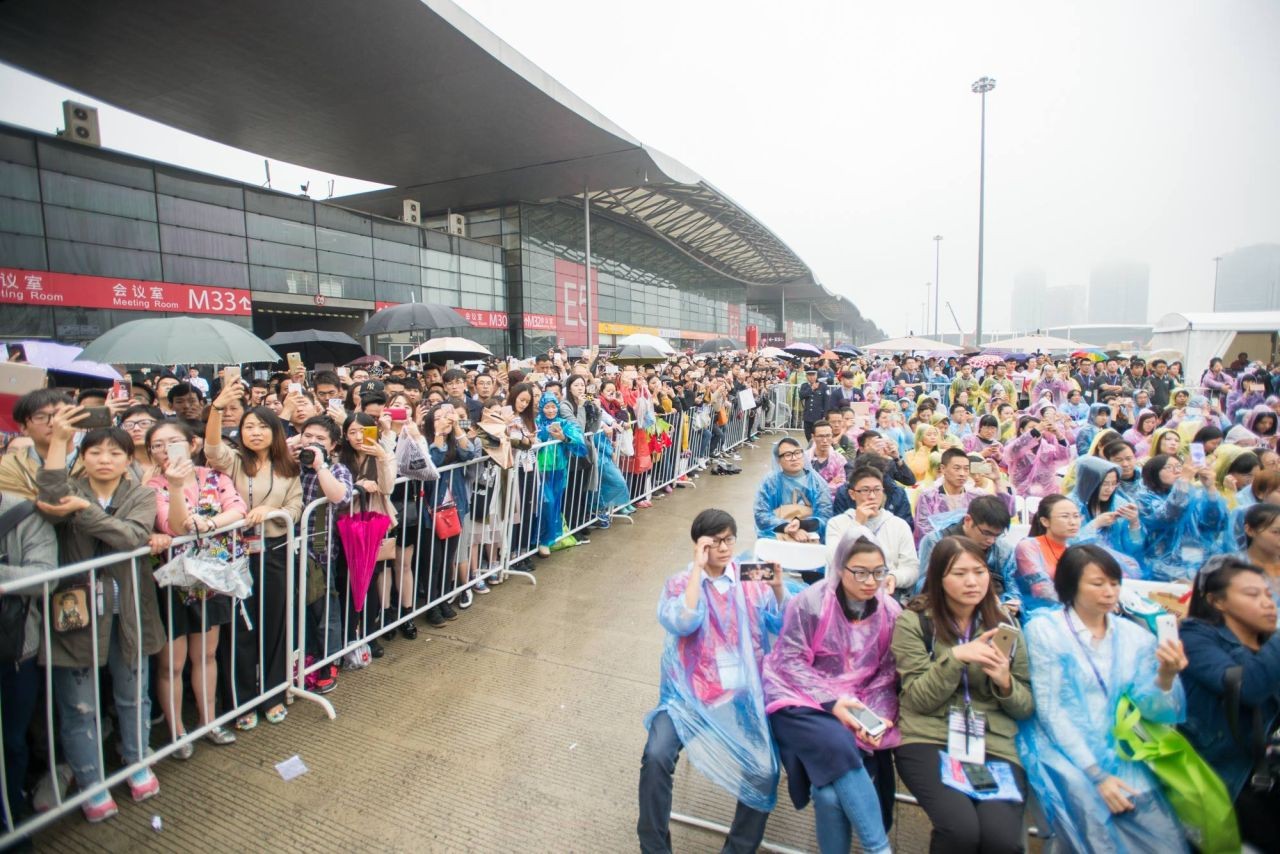 A large crowd, many wearing raincoats and holding umbrellas, gathers outside a modern building on a rainy day. Some people stand behind barricades, while others sit on chairs, all looking toward the front.