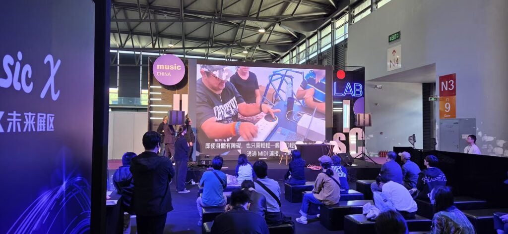 A group of people sit on benches facing a stage with a large screen displaying a demonstration of music equipment at an indoor event called music CHINA. Purple lighting and expo signs are visible in the background.