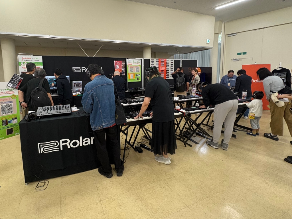 People explore and try out electronic keyboards and synthesizers at a Roland booth inside a spacious indoor event, with various equipment and promotional displays visible.