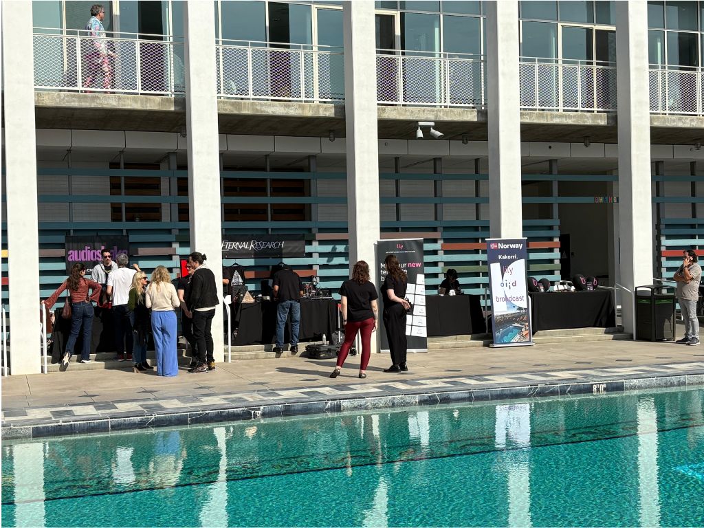 A group of people gather near vendor tables by an outdoor swimming pool, with a modern building featuring large windows and vertical white columns in the background. Some booths have banners and promotional materials displayed.