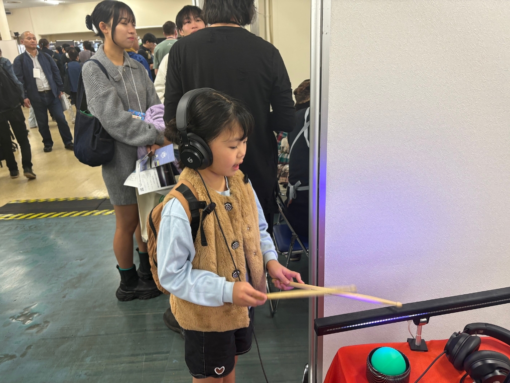 A young girl wearing headphones and holding drumsticks plays an electronic drum pad at an indoor event, with people and booths in the background.