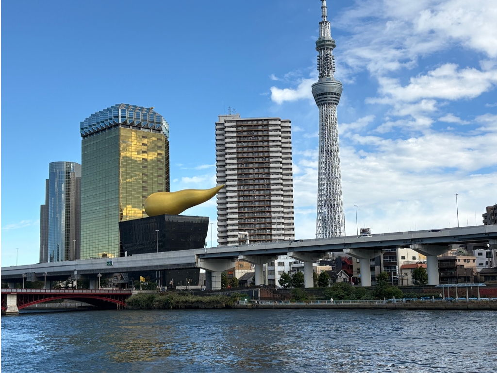 Tokyo cityscape featuring the Asahi Beer Hall with its golden flame sculpture, the Tokyo Skytree tower, modern and high-rise buildings, a bridge, and a river in the foreground under a partly cloudy sky.