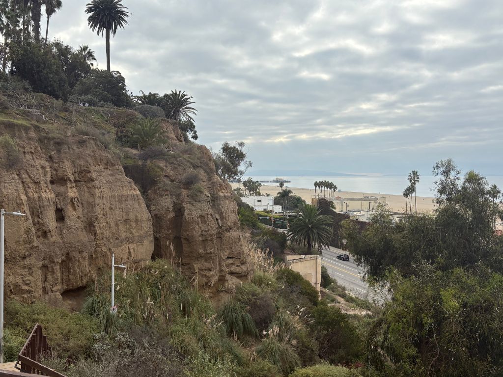 A rocky cliff with palm trees overlooks a road, sandy beach, and the ocean under a cloudy sky. Some buildings and more palm trees are visible near the shoreline in the distance.