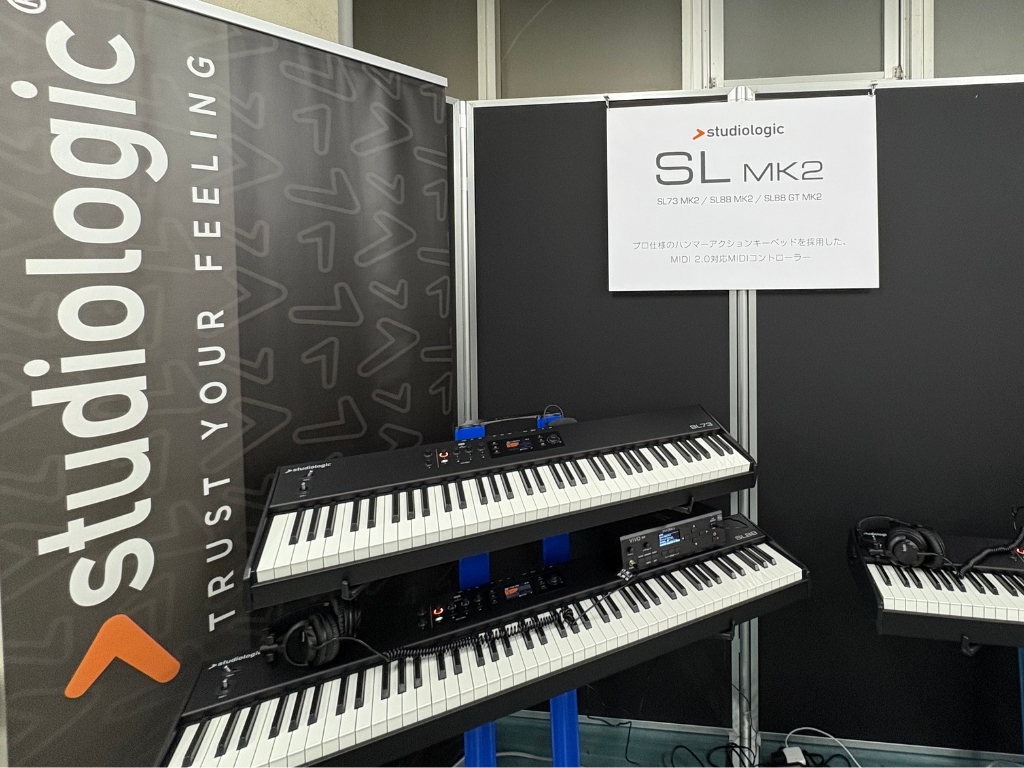 Two Studiologic SL MK2 MIDI keyboard controllers are displayed on stands at an exhibition booth, with a Studiologic banner and sign in the background. Headphones are placed on one of the keyboards.