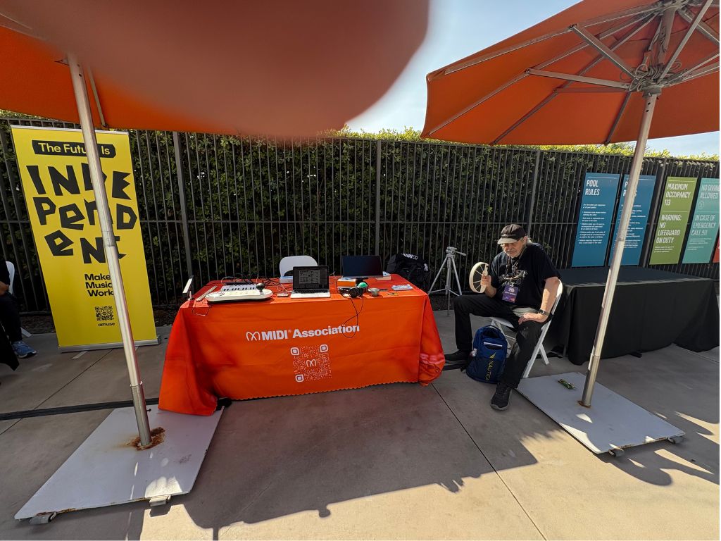 An outdoor booth with a bright orange tablecloth labeled MIDI Association is set up under large umbrellas. A person sits beside the booth, giving a thumbs-up. Colorful banners and informational posters are displayed behind.