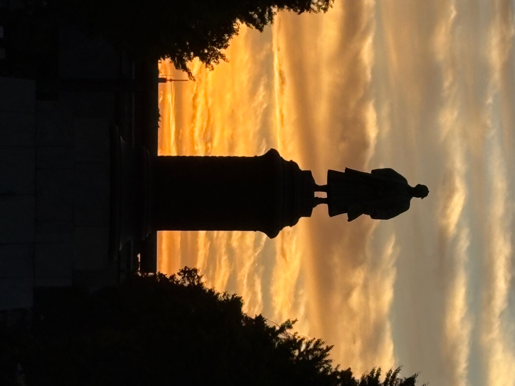 A statue of a standing figure is silhouetted against a dramatic orange and yellow sunset sky, framed by dark trees on either side.