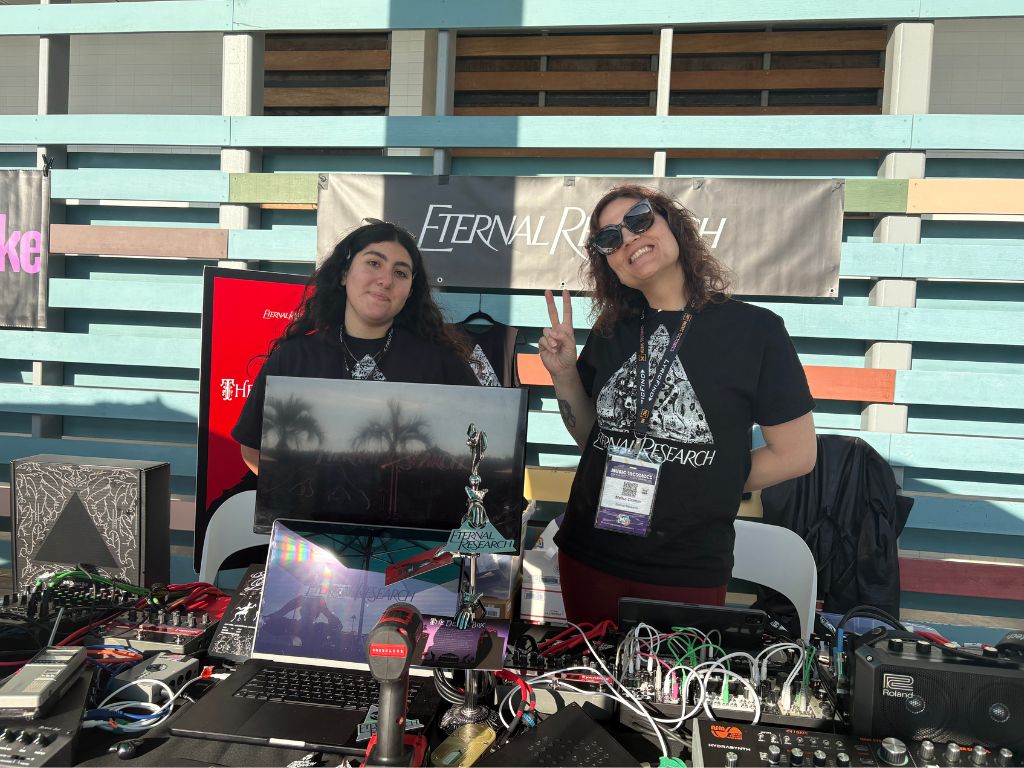 Two women stand behind a table with electronic equipment and laptops, smiling at an outdoor event. One gives a peace sign. Both wear black Eternal Research shirts, with banners and colorful background behind them.