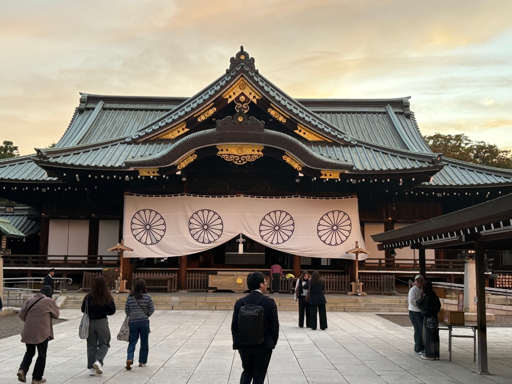 People stand in front of a traditional Japanese temple with ornate roof details and a large white drape featuring circular emblems, under a cloudy evening sky.