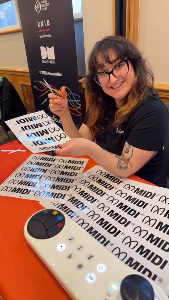 Adi Dickens from Ableton with glasses and a tattoo smiles at a table with MIDI stickers and an Arcana Strum tactile electronic music device. She holds colorful cables, and a banner with logos is visible in the background.