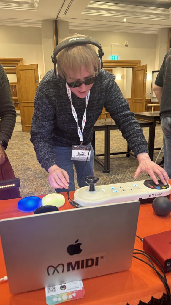 A person wearing headphones and sunglasses uses an accessible DJ controller and laptop at a table with colorful lights. The person is standing and appears focused on the music setup in a conference room.
