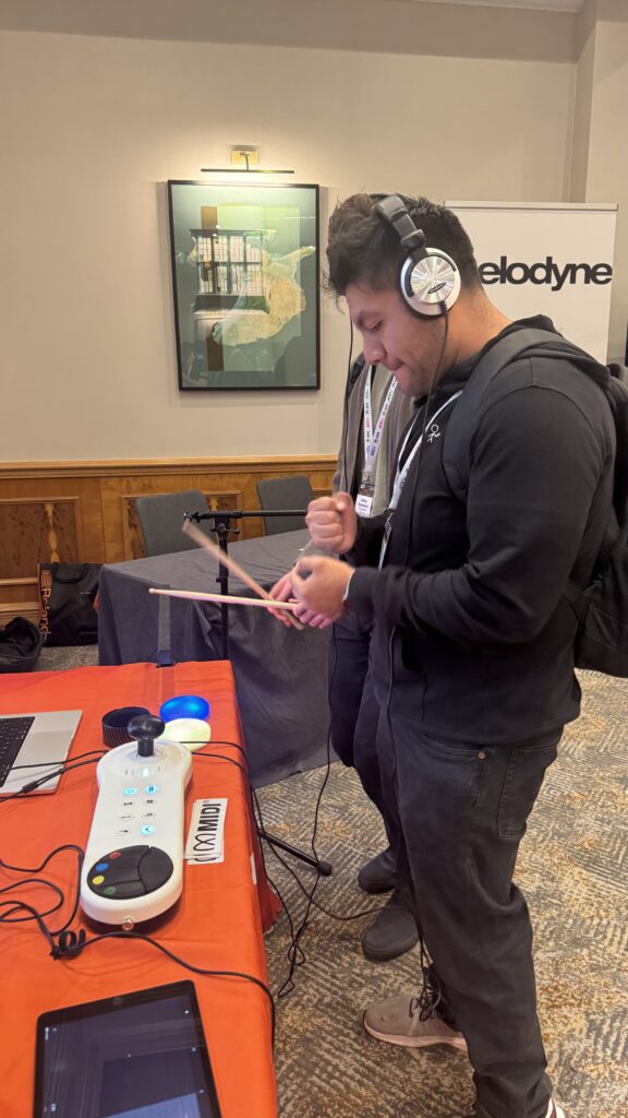 A person wearing headphones holds drumsticks and interacts with an electronic drum pad on a table at an event, with a Velodyne banner and framed artwork visible in the background.