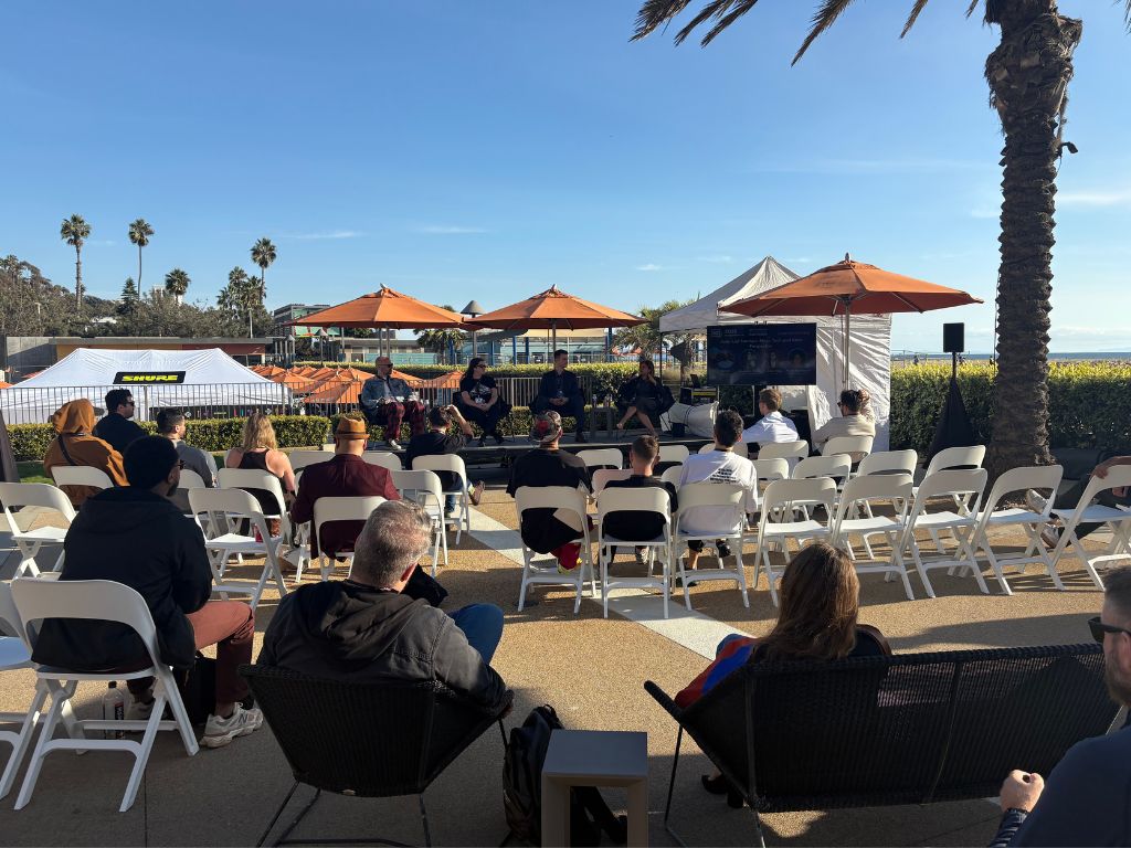 A group of people sit on white chairs facing a panel of speakers under orange umbrellas at an outdoor event near the beach, with palm trees and a blue sky in the background.
