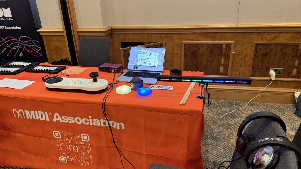 A table with an orange MIDI Association tablecloth displays various electronic music devices, including a MIDI controller, colorful buttons, drumsticks, a laptop, headphones, and a multi-colored LED strip.