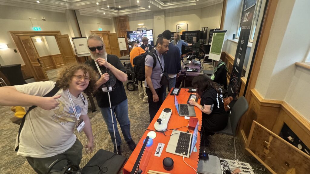 A group of people interact at an indoor event. One person gives a thumbs-up, another uses a white cane, and others stand or sit at a table with laptops and tech equipment. Signs and banners are visible in the background.