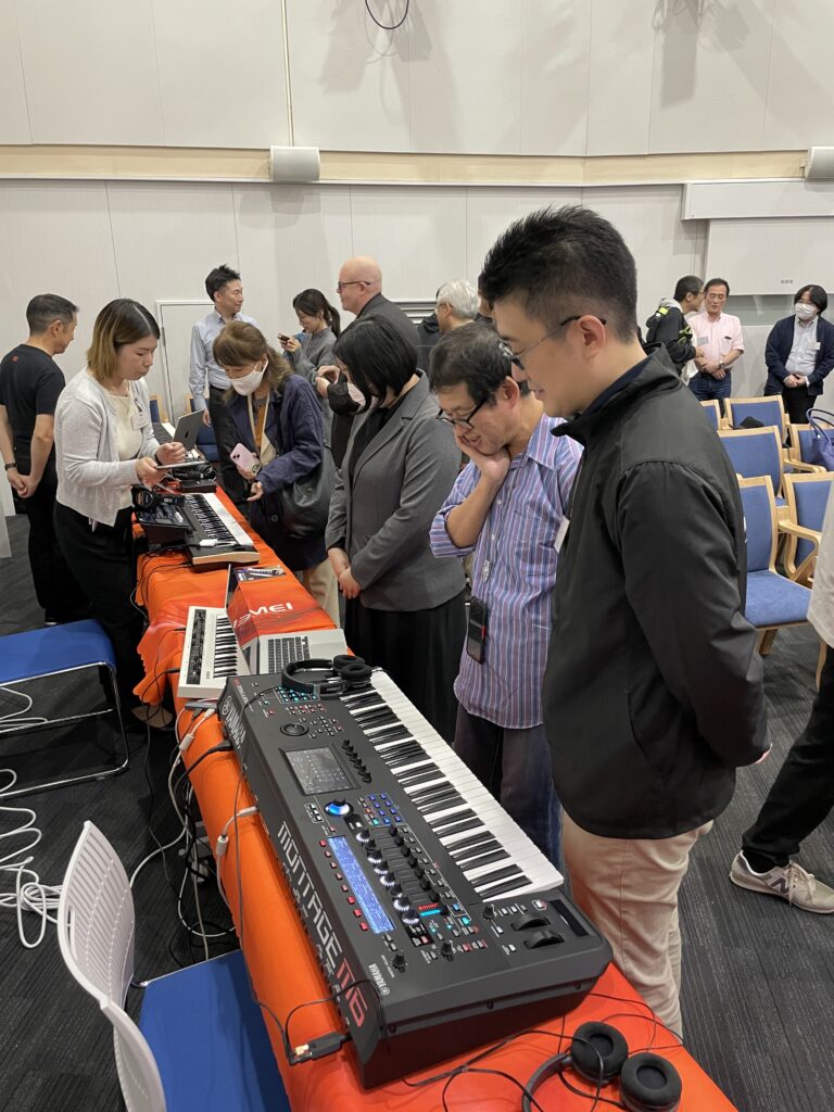 A group of people stand around a table with electronic music equipment, including keyboards and synthesizers, in a well-lit room. Some are observing while others interact with the devices.