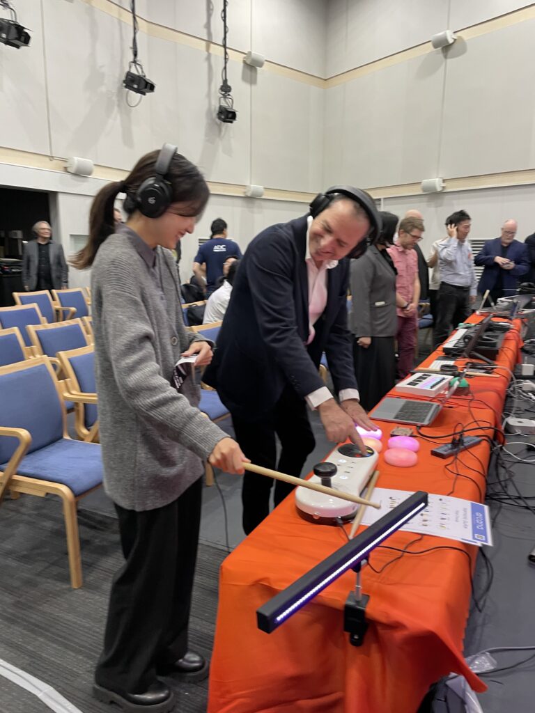 A woman wearing headphones plays an electronic drum pad with drumsticks while a man in a suit, also wearing headphones, smiles and points at the drum pad. They are in a room with chairs, laptops, and other people.